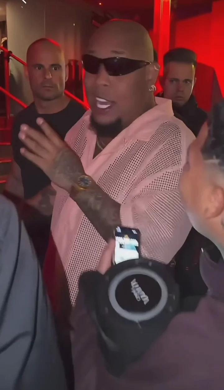 Frame from the supplied Bulin 47 clip: lead performer in sunglasses and a pink shirt, a foreground phone and speaker, and people by red-lit steps